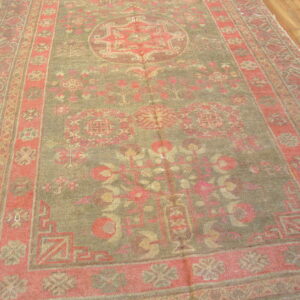 Distressed rug with an olive green field and faded coral border lays on brown hardwood floor.