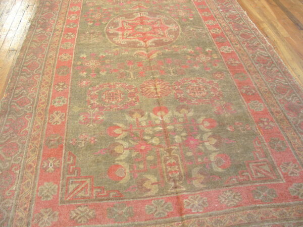Distressed rug with an olive green field and faded coral border lays on brown hardwood floor.