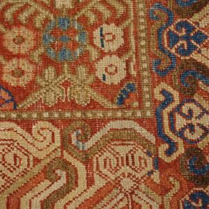 Knotted rug detail in rusty red, beige, brown, and blue, showing complex floral and scrolling patterns.