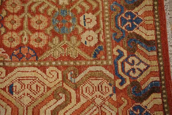 Knotted rug detail in rusty red, beige, brown, and blue, showing complex floral and scrolling patterns.