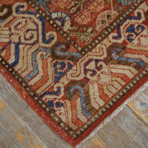 Close-up of a low-pile rug showing red, blue, tan, and brown traditional patterns on rustic wood.