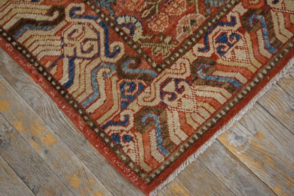 Close-up of a low-pile rug showing red, blue, tan, and brown traditional patterns on rustic wood.