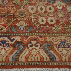 Textured rug close-up showing red floral motifs, geometric blue borders, and short white fringe.