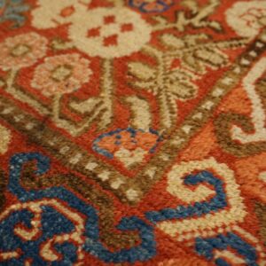 Textured, close-up view of a rug featuring red, tan, blue, and brown colors in a traditional pattern.
