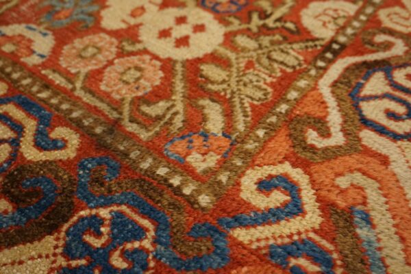 Textured, close-up view of a rug featuring red, tan, blue, and brown colors in a traditional pattern.