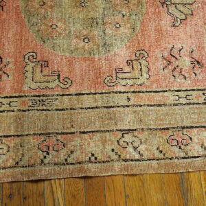 Distressed pink and tan rug featuring geometric motifs and an olive medallion on wood plank flooring.