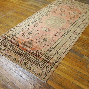 Faded pink runner rug with dark geometric borders sits on a distressed, warm brown hardwood floor.