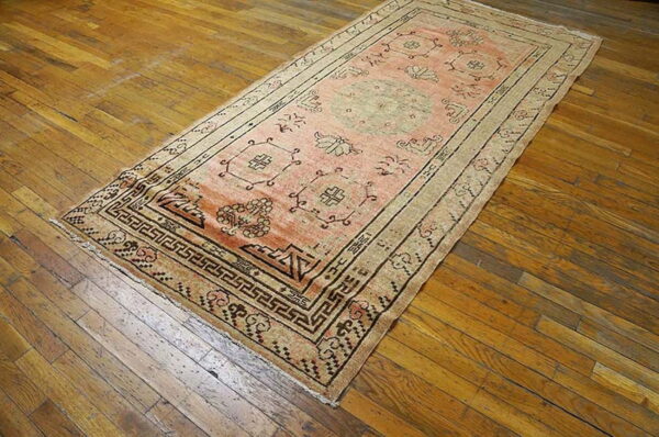 Faded pink runner rug with dark geometric borders sits on a distressed, warm brown hardwood floor.