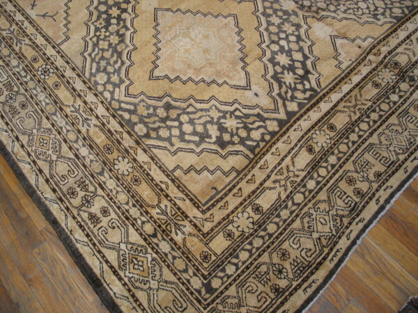 A close-up of a tan and dark brown geometric rug corner lying on medium brown wood flooring.