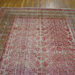 Distressed beige and red patterned rug with multiple borders laid over brown hardwood flooring.