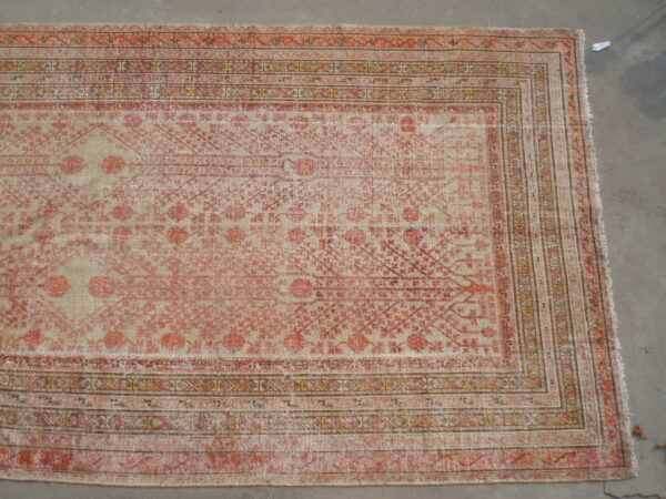 Distressed rug with faded salmon and beige geometric patterns and multi-stripe borders on a gray concrete floor.