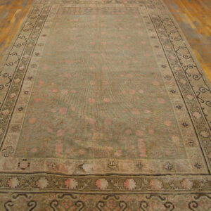 Low-pile rug in muted olive green and faded pink with detailed floral borders rests on wood plank flooring.