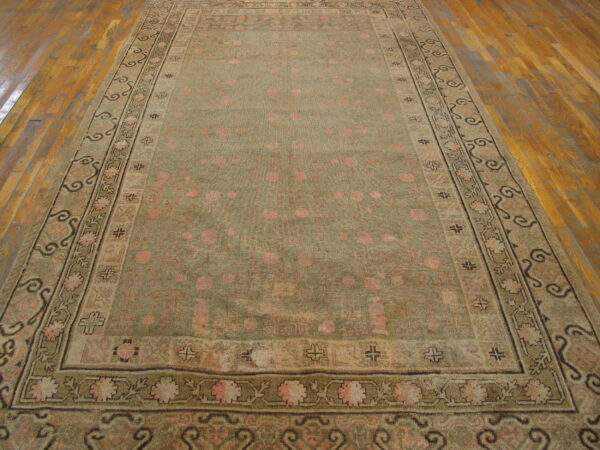 Low-pile rug in muted olive green and faded pink with detailed floral borders rests on wood plank flooring.