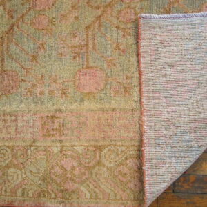 Folded corner of a green and pink patterned rug shows its woven backing over worn wood flooring.