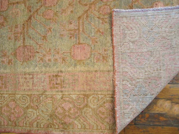Folded corner of a green and pink patterned rug shows its woven backing over worn wood flooring.