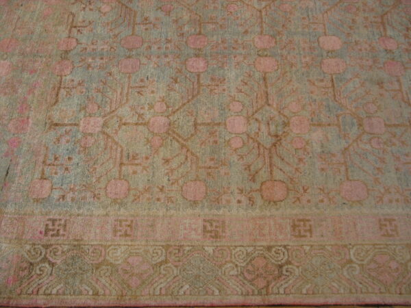 Low-pile rug with geometric pink, tan, and pale blue patterns, including stylized medallions and layered borders.