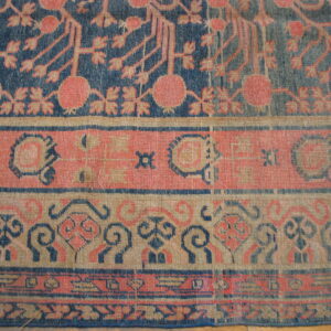 Worn, low-pile rug section displaying intricate navy, coral, and tan patterns on plank hardwood flooring.