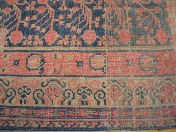 Worn, low-pile rug section displaying intricate navy, coral, and tan patterns on plank hardwood flooring.