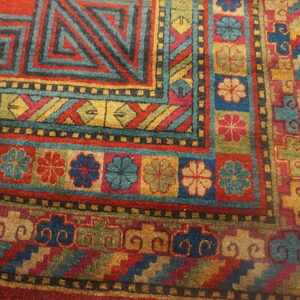 Geometric and floral patterned rug corner features red, teal, gold, and olive colors on distressed wood flooring.