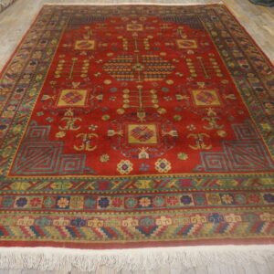 Large red rug featuring intricate geometric and stylized botanical patterns in gold, green, and blue on wood planks.