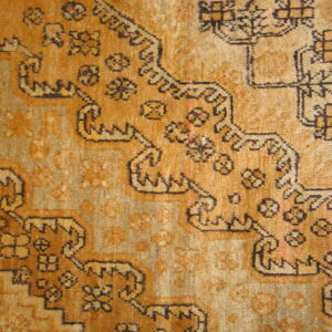 Textured rug close-up showing golden-brown and tan colors with complex stepped geometric designs.