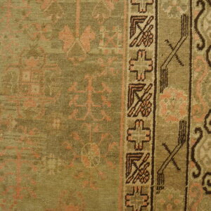 Detail of a low-pile olive rug with faded peach motifs and a dark geometric border pattern.