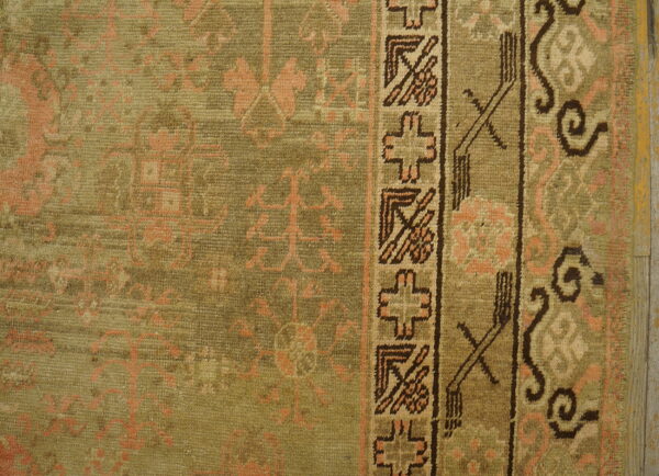 Detail of a low-pile olive rug with faded peach motifs and a dark geometric border pattern.