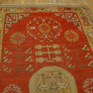 Rusty orange rug featuring mustard yellow borders and geometric patterns rests on distressed wood planks.