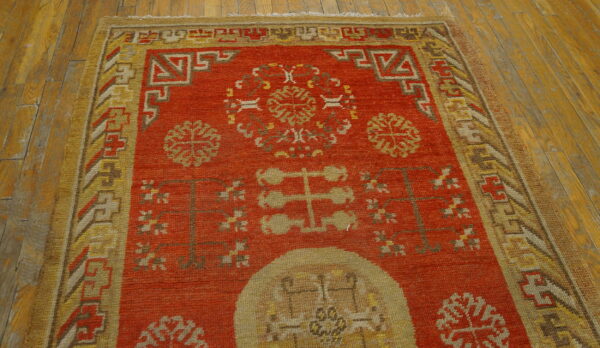 Rusty orange rug featuring mustard yellow borders and geometric patterns rests on distressed wood planks.