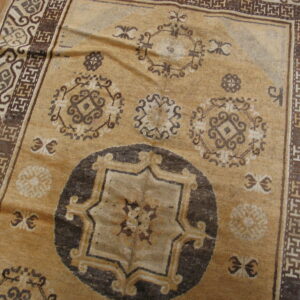 Tan rug with dark brown geometric medallions and complex borders resting on light wood flooring.