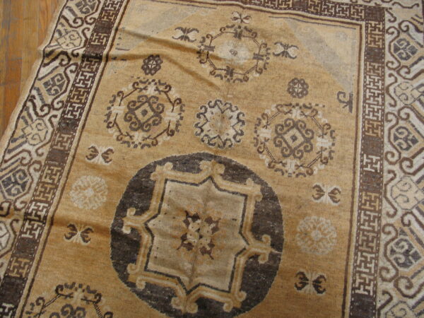 Tan rug with dark brown geometric medallions and complex borders resting on light wood flooring.