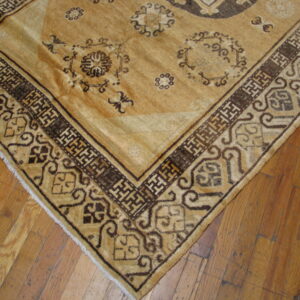 Tan rug corner showing a dark brown fretwork border and floral medallions on rustic wooden plank flooring.