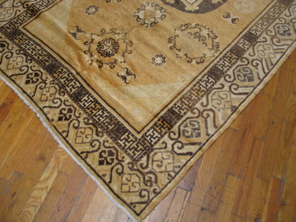 Tan rug corner showing a dark brown fretwork border and floral medallions on rustic wooden plank flooring.