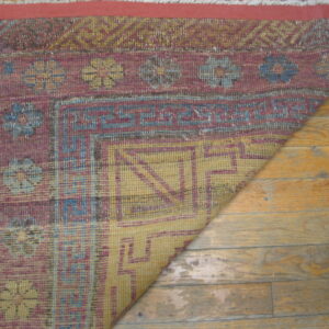 Low-pile rug in maroon, yellow, and blue geometric and floral patterns lays partially folded on hardwood floor.