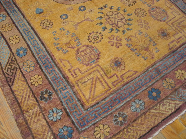 Ochre rug featuring complex geometric borders and floral motifs in purple and light blue on a hardwood floor.