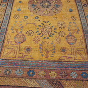 Gold field rug with blue geometric key border and floral maroon border sits on wood flooring.