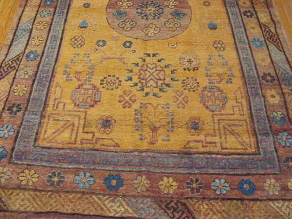 Gold field rug with blue geometric key border and floral maroon border sits on wood flooring.