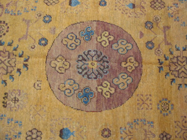 Golden yellow low-pile rug showing a round purple center motif with scrolling blue and tan designs.