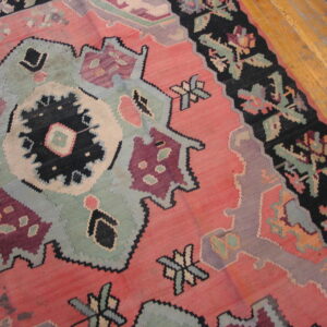 Flatwoven rug featuring large stylized motifs in pink, black, dusty blue, and maroon on distressed wood flooring.