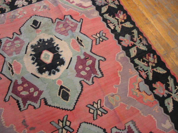Flatwoven rug featuring large stylized motifs in pink, black, dusty blue, and maroon on distressed wood flooring.