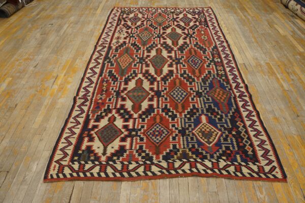 Low pile rug with red, cream, navy, and black geometric patterns on worn light hardwood flooring.