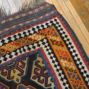 Flatwoven rug with geometric patterns in blue, orange, and red, showing long fringe on wood planks.