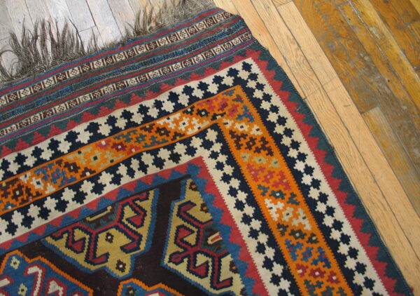 Flatwoven rug with geometric patterns in blue, orange, and red, showing long fringe on wood planks.