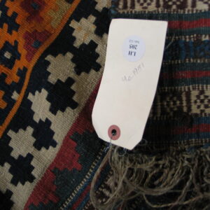 Multicolored geometric flatweave rug in orange, navy, and tan, showing fringe and an inventory tag.