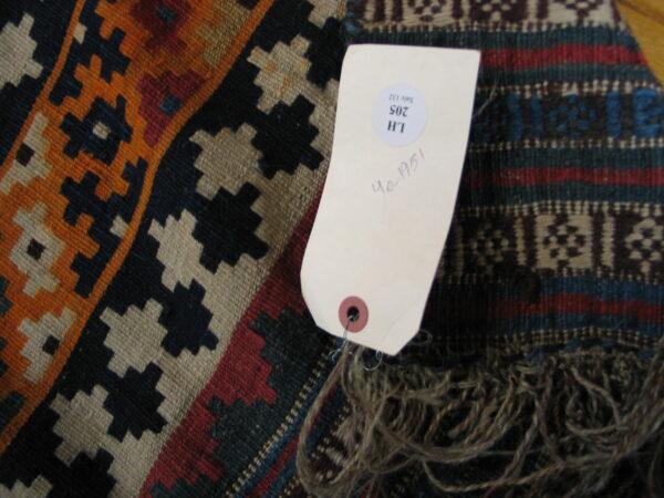 Multicolored geometric flatweave rug in orange, navy, and tan, showing fringe and an inventory tag.