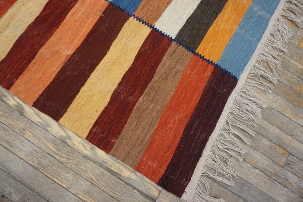 A flatwoven patchwork rug featuring wide stripes in burgundy, orange, tan, and blue rests on distressed wooden floors.