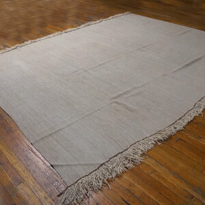 A solid light gray flatweave rug with long fringe rests on distressed brown hardwood floorboards.