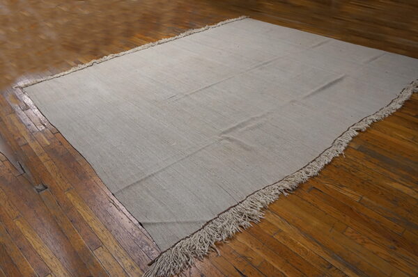 A solid light gray flatweave rug with long fringe rests on distressed brown hardwood floorboards.