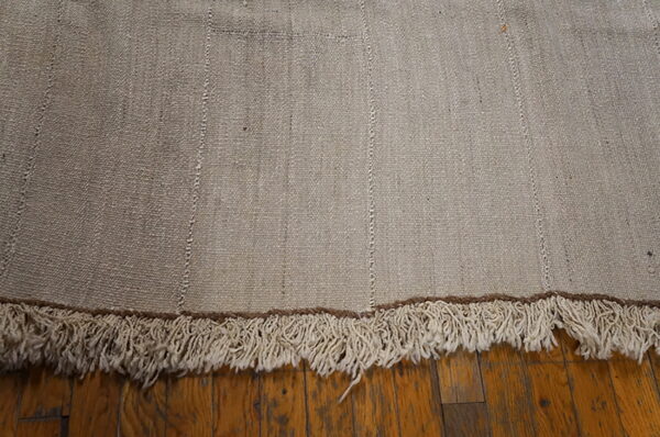 A light brown flatweave rug with stitched vertical accents and shaggy cream fringe rests on wood planks.