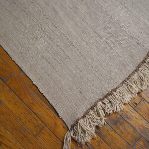 A coarse, flatwoven, light beige rug with subtle stripes and thick fringe rests diagonally on distressed wooden floors.